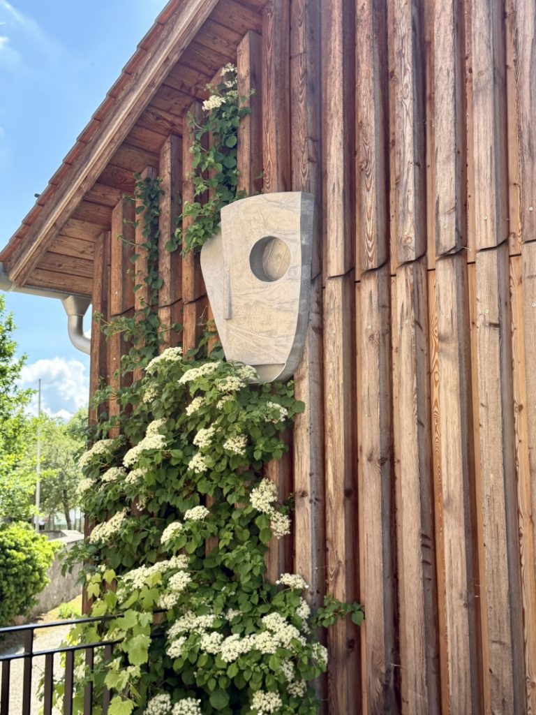 Holzhaus mit Blumenranken, darin eine weiße Skulptur
